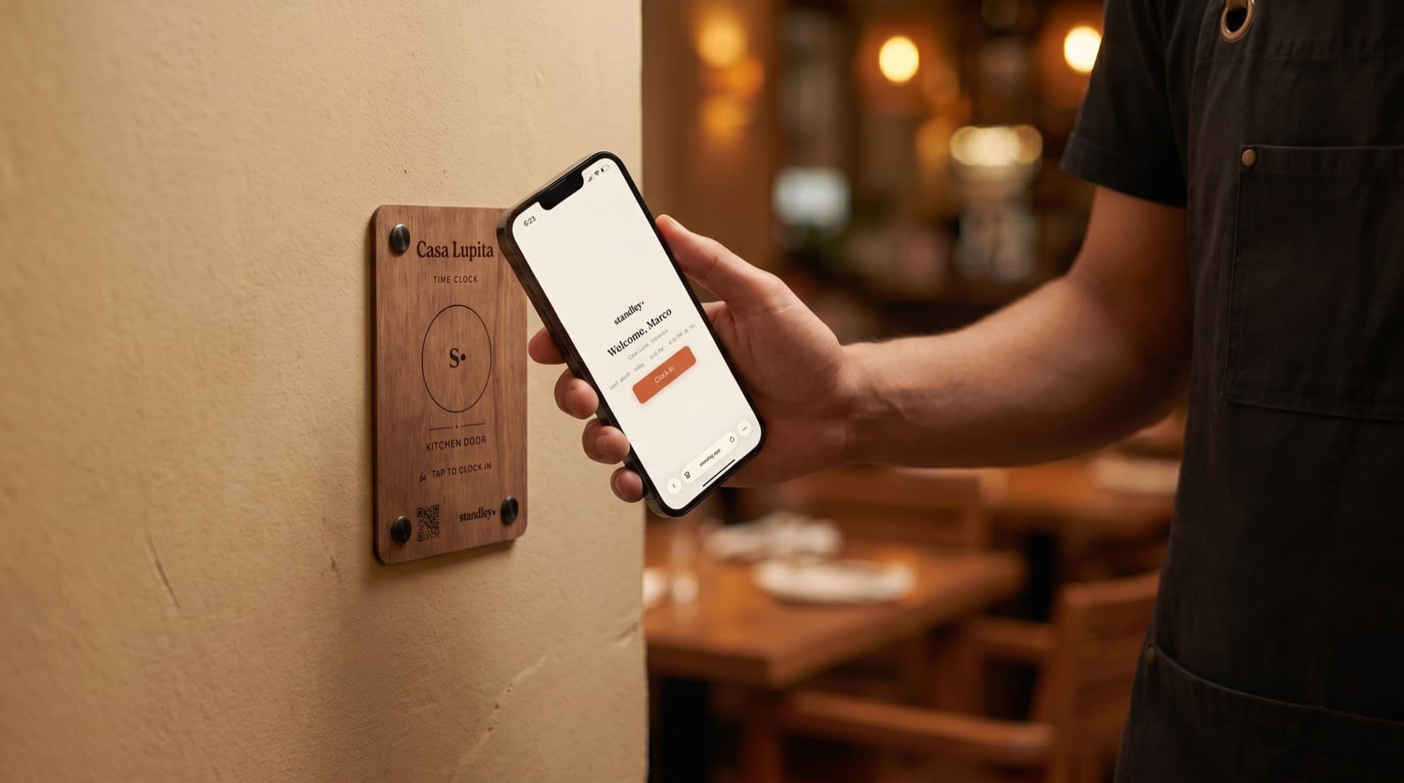A cook taps their phone on the engraved Casa Lupita clock-in station by the kitchen door — the standley app greets them 'Welcome, Marco' with a Clock in button